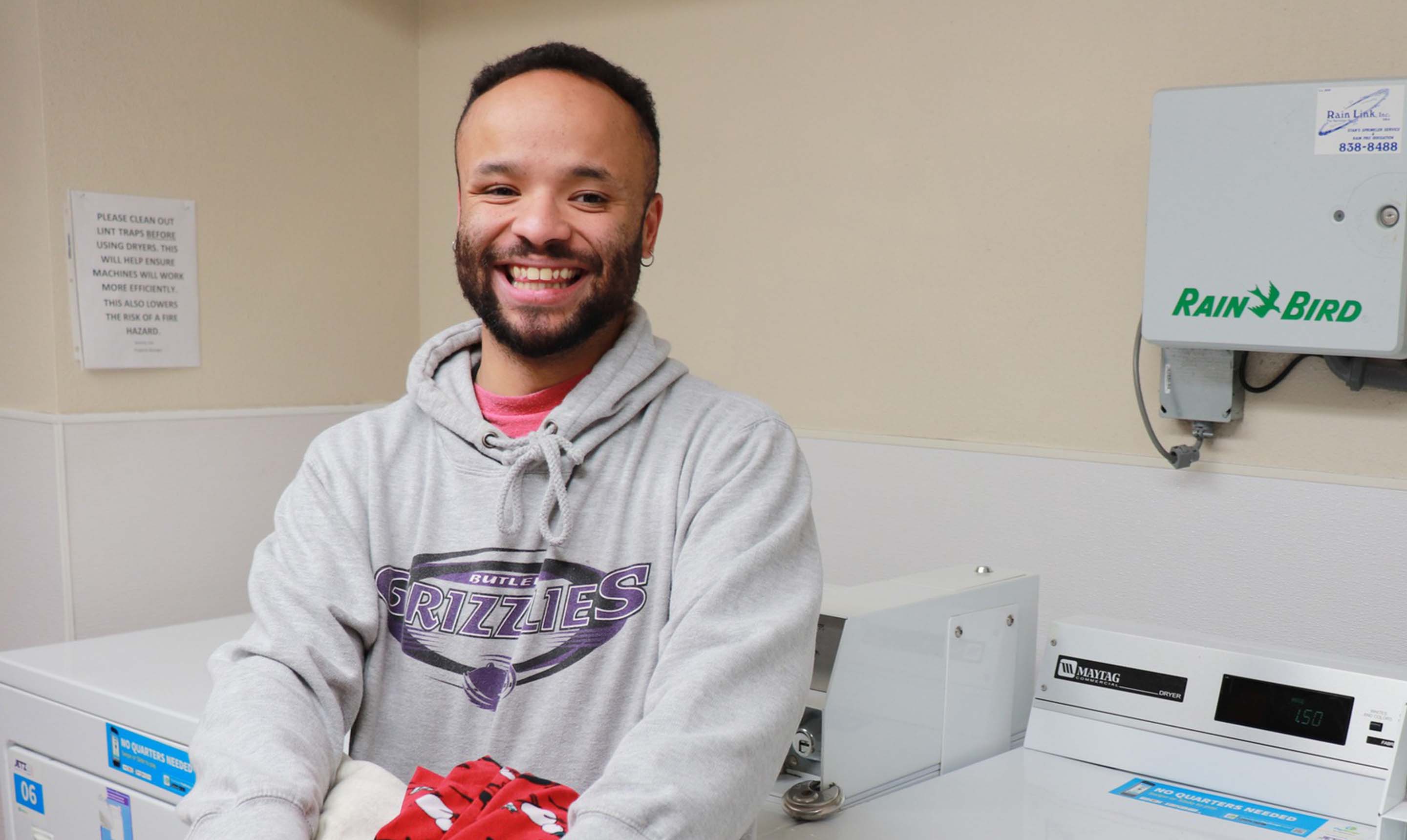 A GILA participant is in the laundry room at The Timbers, holding a basket of clean clothes and smiling towards the camera.