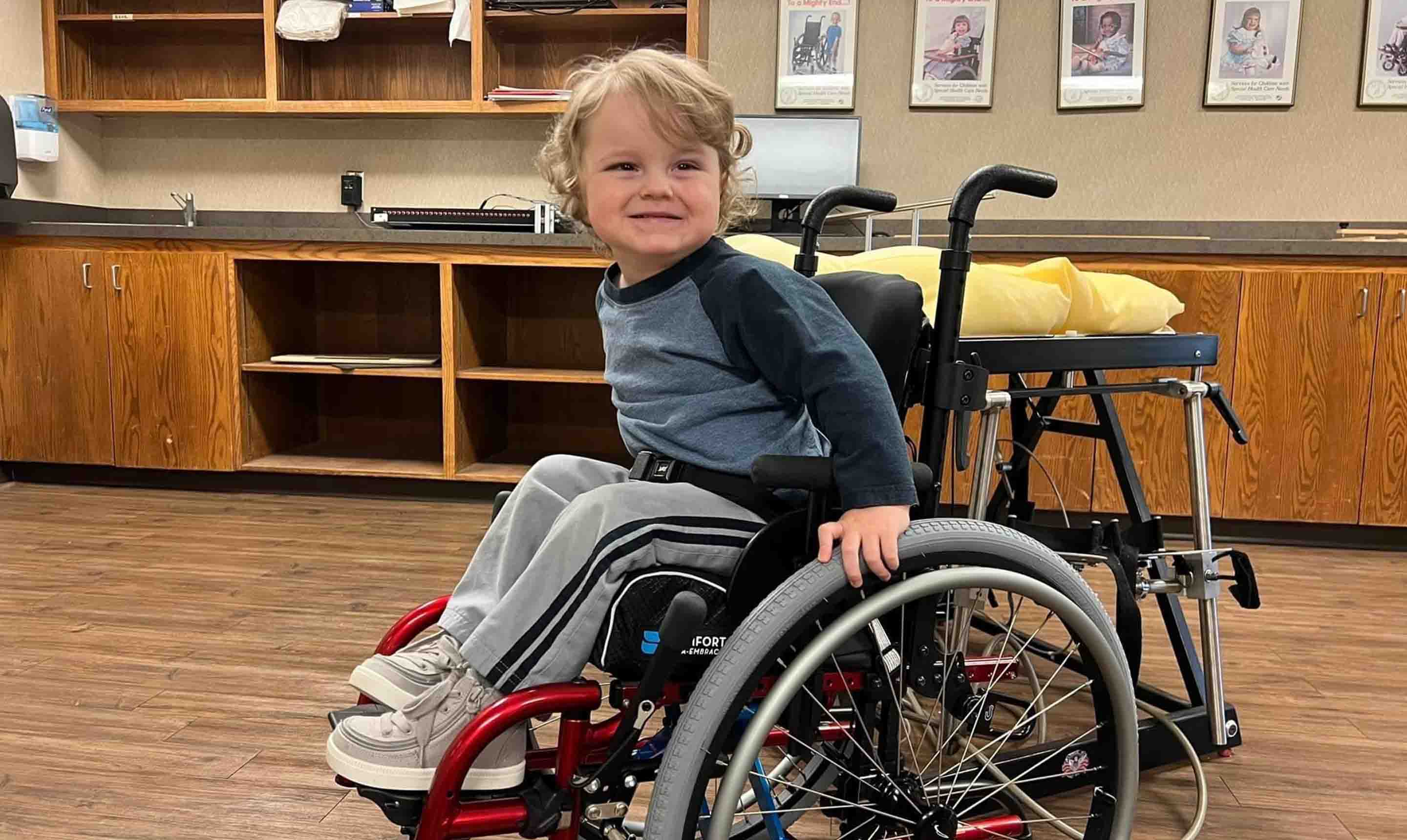 A young Wheelchair Clinic client is inside an exam room, seated in his red manual wheelchair and smiling towards the camera.