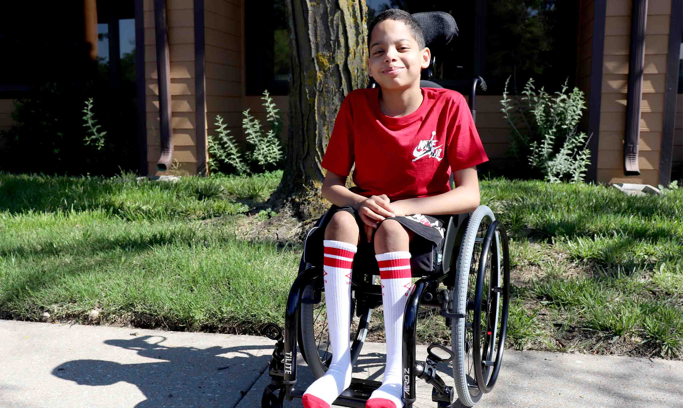 A young CPRF Wheelchair Clinic client is seated outside of the building in his manual chair, wearing a red t-shirt, shorts, and red and white tube socks.