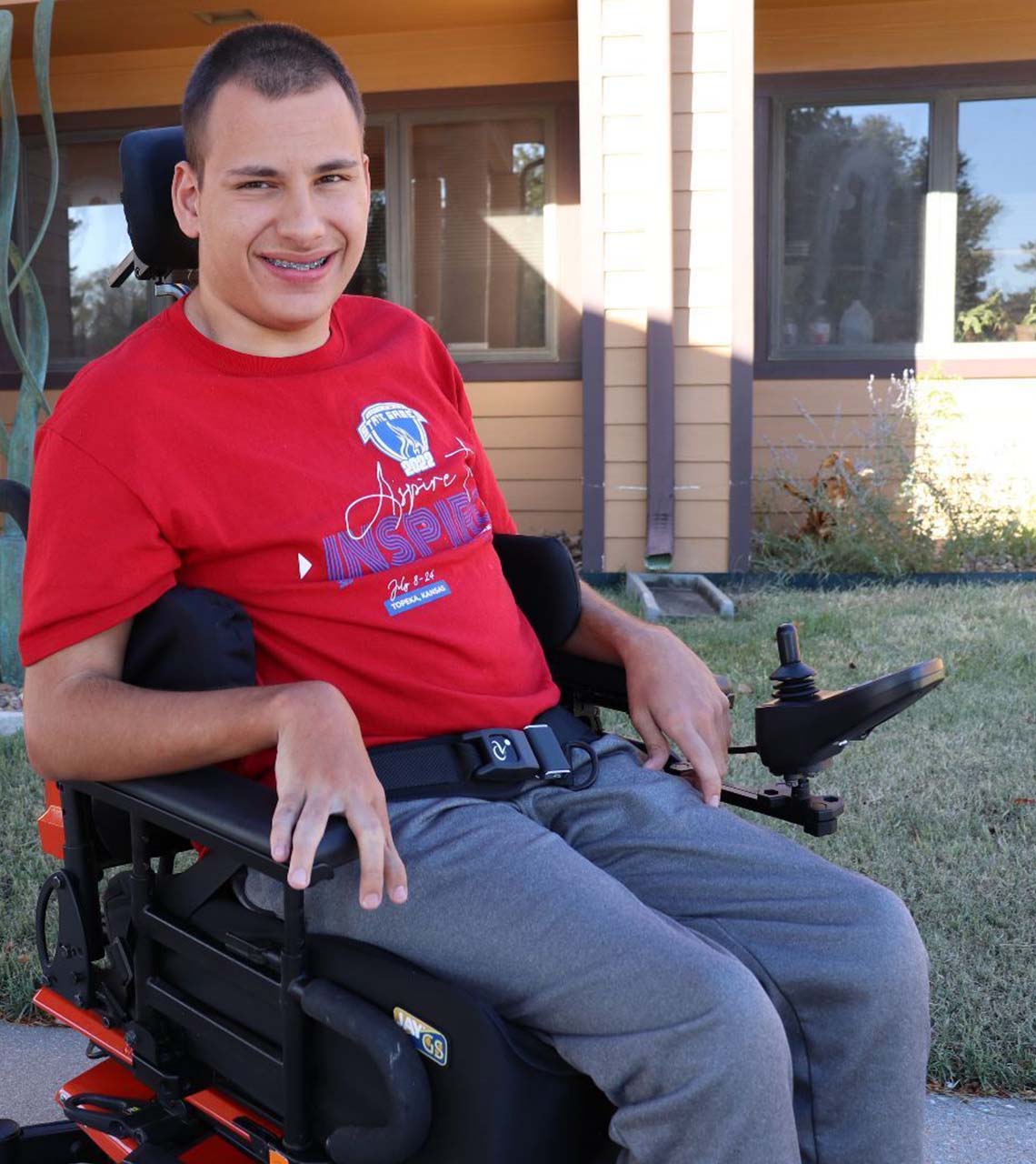 Photo of a Wheelchair Clinic client seated outside of the building. He is in an orange powerchair, wearing a red t-shirt and jeans.